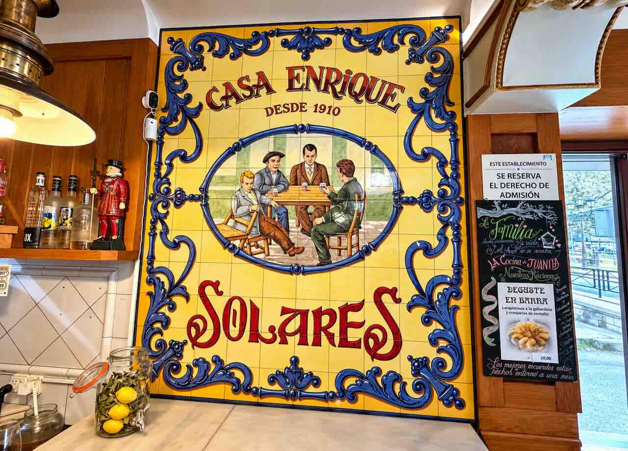 Panel de azulejos amarillos y azules con una escena de sobremesa y la inscripción «Casa Enrique, desde 1910, Solares».